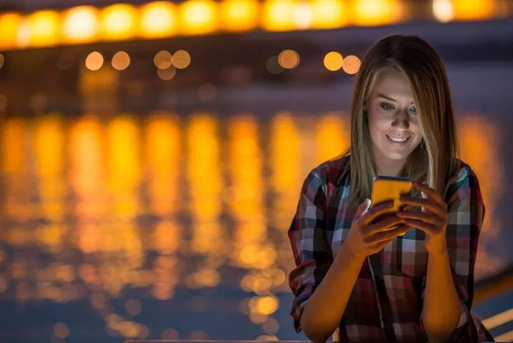 Eine Person in kariertem Hemd lächelt auf ihr leuchtendes Smartphone blickend. Im Hintergrund sind unscharfe orange Lichter und Wasserspiegelungen zu sehen.
