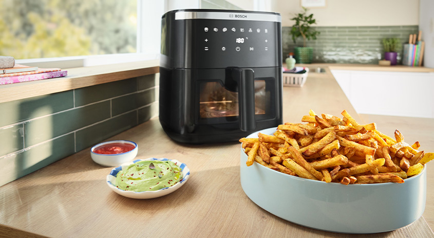  Schüssel mit goldgelb gebackenen Pommes frites auf einer Küchenarbeitsplatte vor einer schwarzen Heißluftfritteuse, daneben kleine Schälchen mit Ketchup und grünem Dip.