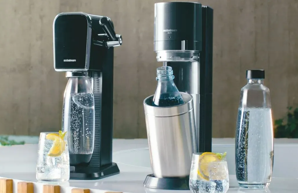 Zwei schwarze SodaStream-Geräte, jeweils mit Flaschen. Drei Flaschen und zwei Gläser mit Sprudelwasser, Eis und Zitrone auf weißer Fläche.