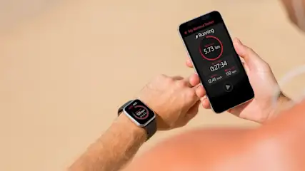 Eine Hand hält ein Smartphone mit Lauf-App (5.73 km, 132 bpm). Eine Smartwatch am Handgelenk zeigt 120 bpm. Heller Hintergrund.