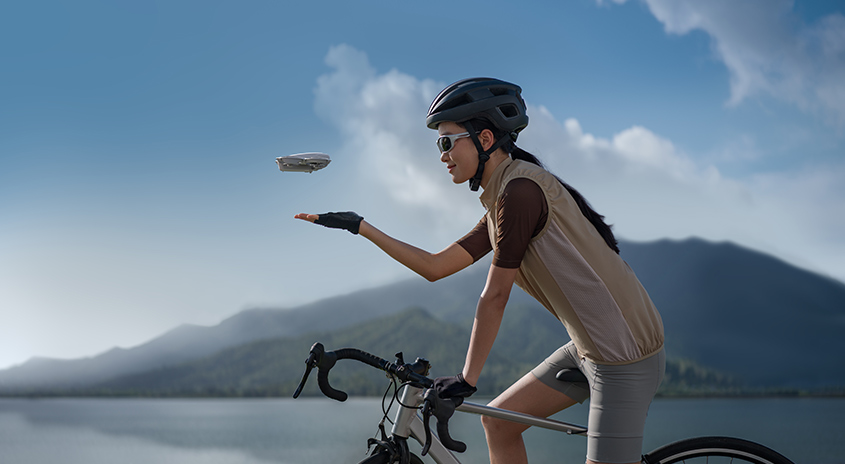 Radfahrer mit Helm hält Drohne vor Bergen und blauem Himmel in der Hand.