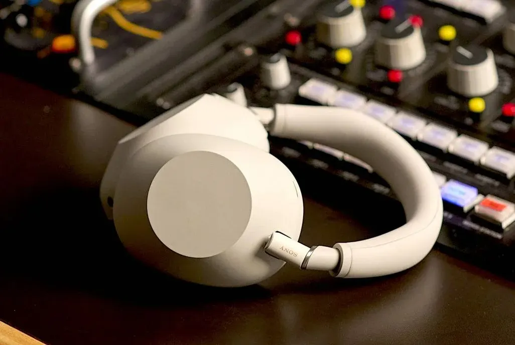 Weiße Sony-Kopfhörer liegen auf einer dunklen Oberfläche vor unscharfem Musikproduktions-Equipment mit Reglern, bunten Kontrollleuchten (rot, gelb, blau) und Kabeln im Hintergrund.