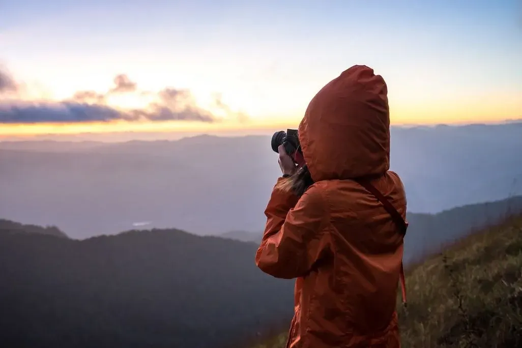 Eine Person in oranger Kapuzenjacke fotografiert mit einer schwarzen Kamera eine weite, neblige Berglandschaft vor blau-orangem Dämmerungshimmel.