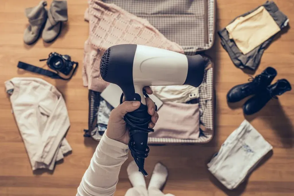 Eine Hand hält einen schwarz-weißen Föhn über einem offenen Koffer. Kleidung, Schuhe und eine Kamera liegen auf Holzboden, bereit zum Packen.