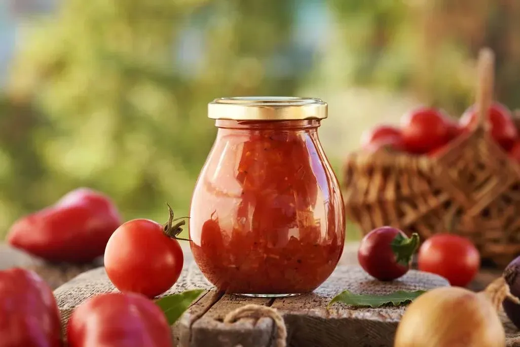 Einmachglas mit hausgemachter Tomatensauce auf Holzbrett, umgeben von frischen Tomaten, roter Paprika und Zwiebeln vor grünem Hintergrund.