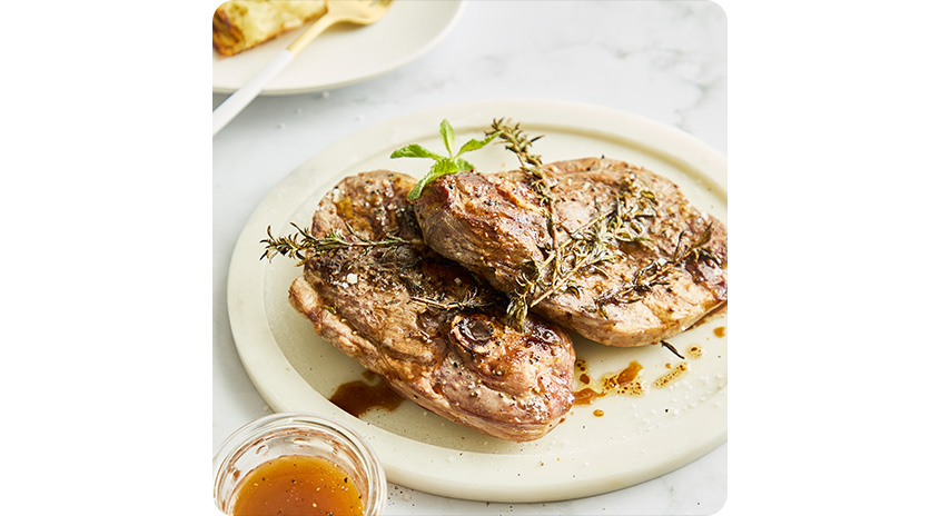  Zwei gebratene Fleischstücke auf hellem Teller, mit frischem Rosmarin garniert, daneben kleine Schale mit brauner Sauce auf hellem Marmortisch.