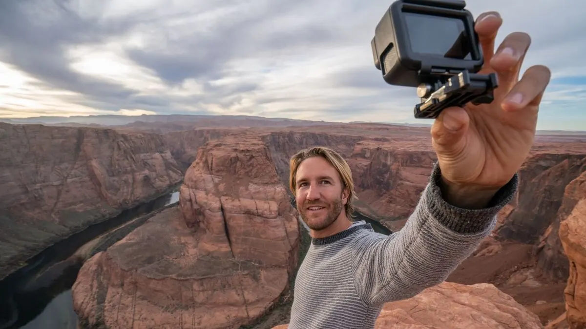 Person im grauen Pullover macht ein Selfie mit schwarzer Actionkamera vor weitem, roten Canyon mit Fluss unter bewölktem Himmel.