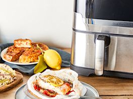 Airfryer, davor stehen zwei Teller mit Fisch mit Zitronenschreibe darauf und auf dem zweiten Teller liegt Gebäck und eine Zitrone davor