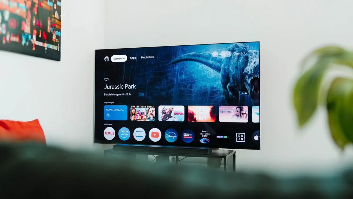 Smart-TV zeigt "Jurassic Park" und Streaming-Apps auf dunklem Ständer. Sofa, rote Kissen und grüne Pflanze im Vordergrund.