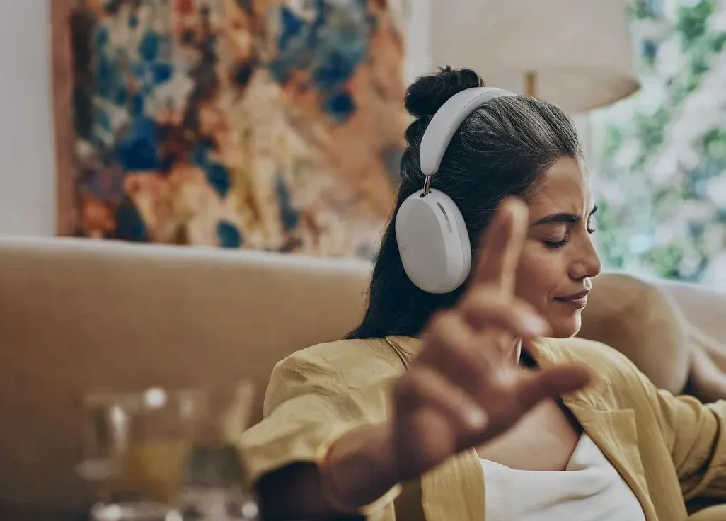 Lächelnde Frau mit weißen Kopfhörern sitzt auf einem Sofa und genießt Musik, Hand ausgestreckt in entspannter Pose.