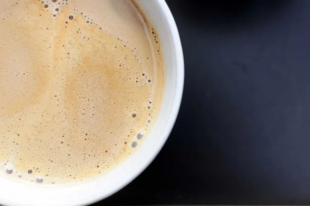 Weiße Tasse mit hellem, schaumigem Kaffee, bedeckt mit Bläschen, auf dunklem Hintergrund.
