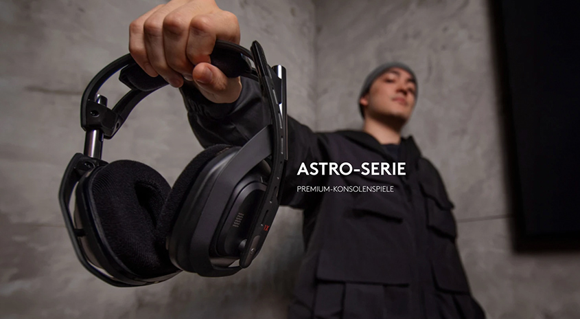 Eine Hand hält ein schwarzes Gaming-Headset vor einer grauen Wand, im Hintergrund ist eine Person zu sehen.