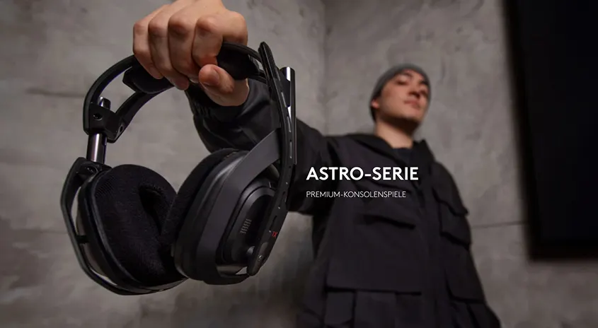 Eine Hand hält ein schwarzes Gaming-Headset vor einer grauen Wand, im Hintergrund ist eine Person zu sehen.