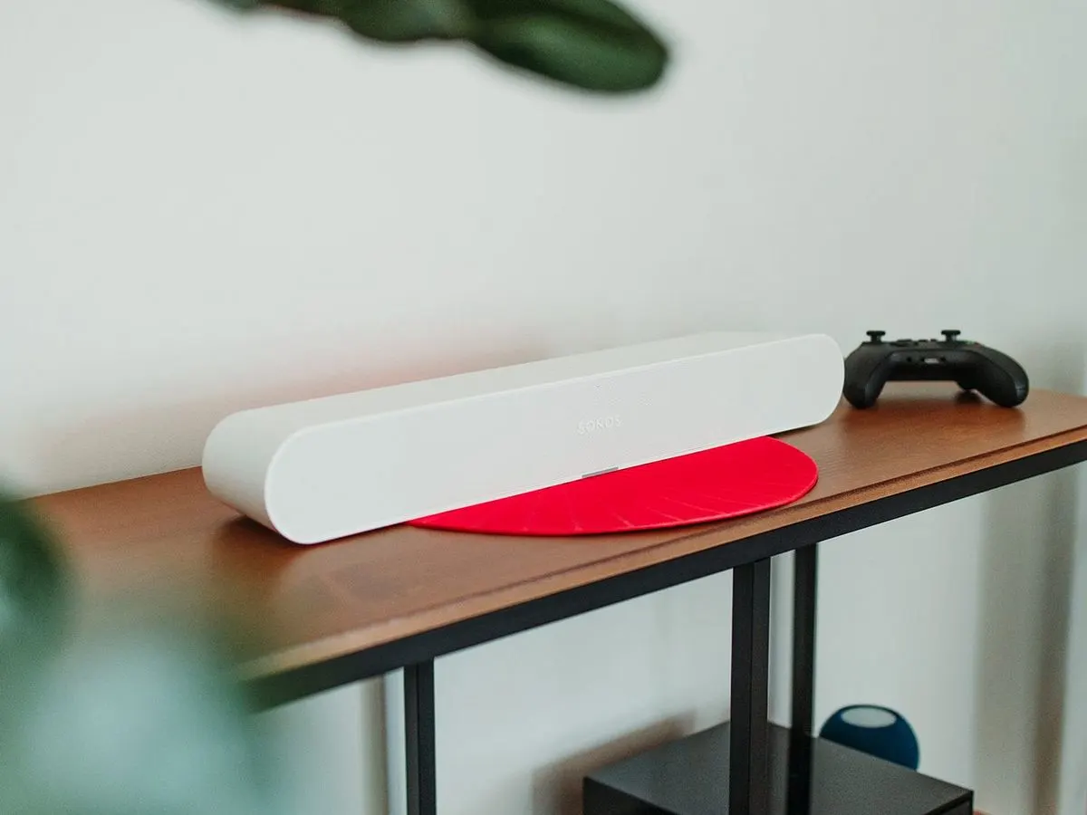 Weißer Sonos-Lautsprecher auf roter Matte, daneben schwarzer Gamecontroller auf Holzregal. Weißer Hintergrund, grüne Blätter.