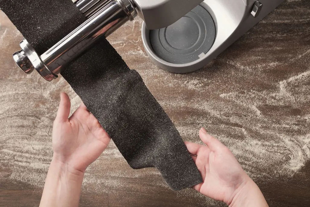 Hände führen dunklen, bemehlten Nudelteig durch eine silberne Nudelmaschine auf einem mehlbestäubten braunen Holztisch mit grauem Mixer.
