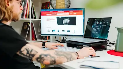 Ein Mann mit Brille tippt auf weißer Tastatur, nutzt Maus an Schreibtisch. Monitor zeigt MediaMarkt-Website, Laptop Symbole. Grüne Kanne rechts, farbige Alben im Regal.