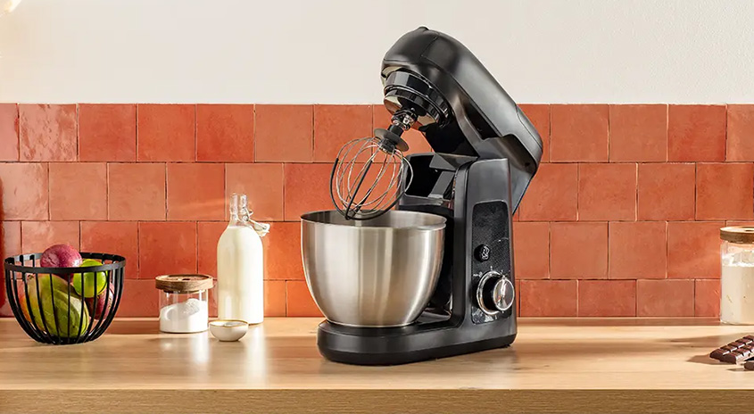 Eine schwarze Küchenmaschine mit Edelstahlschüssel steht auf einer Holztheke vor einer Ziegelwand. Daneben stehen Obst, Milch und Zucker.