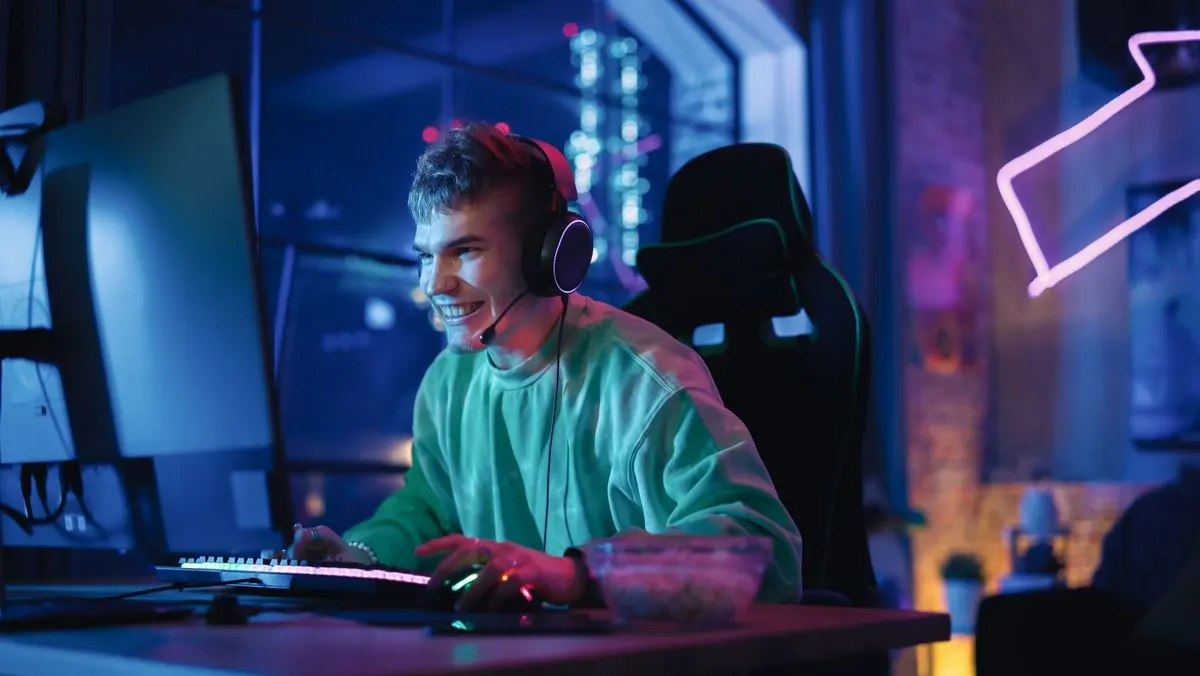 Lächelnder Spieler mit Headset am blauen Monitor, tippt auf beleuchteter Tastatur. Gaming-Stuhl, Snackschale, lila Neonlicht.