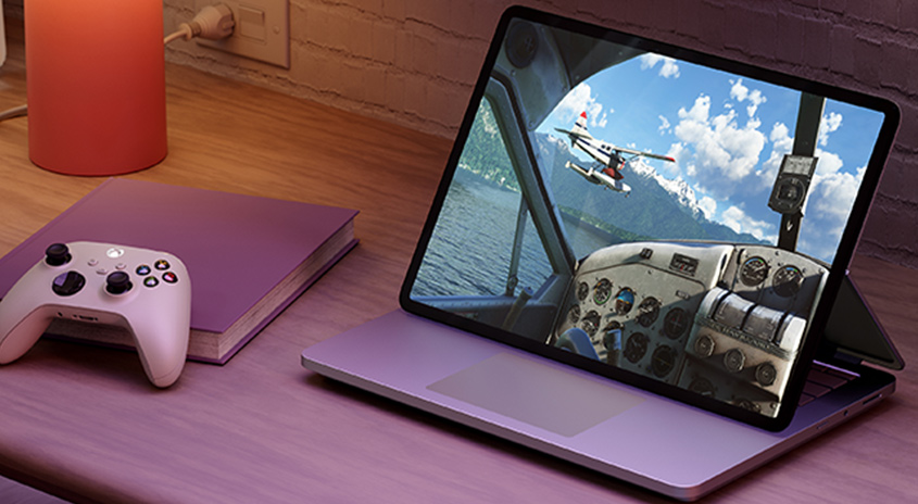 Ein Laptop zeigt ein Flugsimulator-Spiel mit einem Flugzeug über einem See und Bergen. Ein Controller und ein Buch liegen daneben.