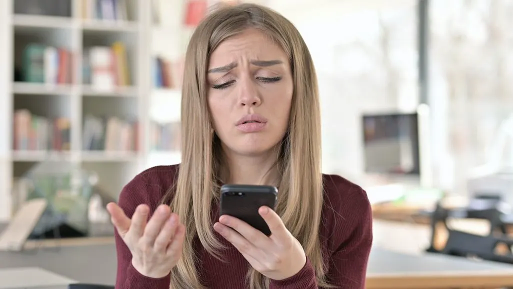 Frau schaut genervt auf Smartphone und gestikuliert mit der freien Hand