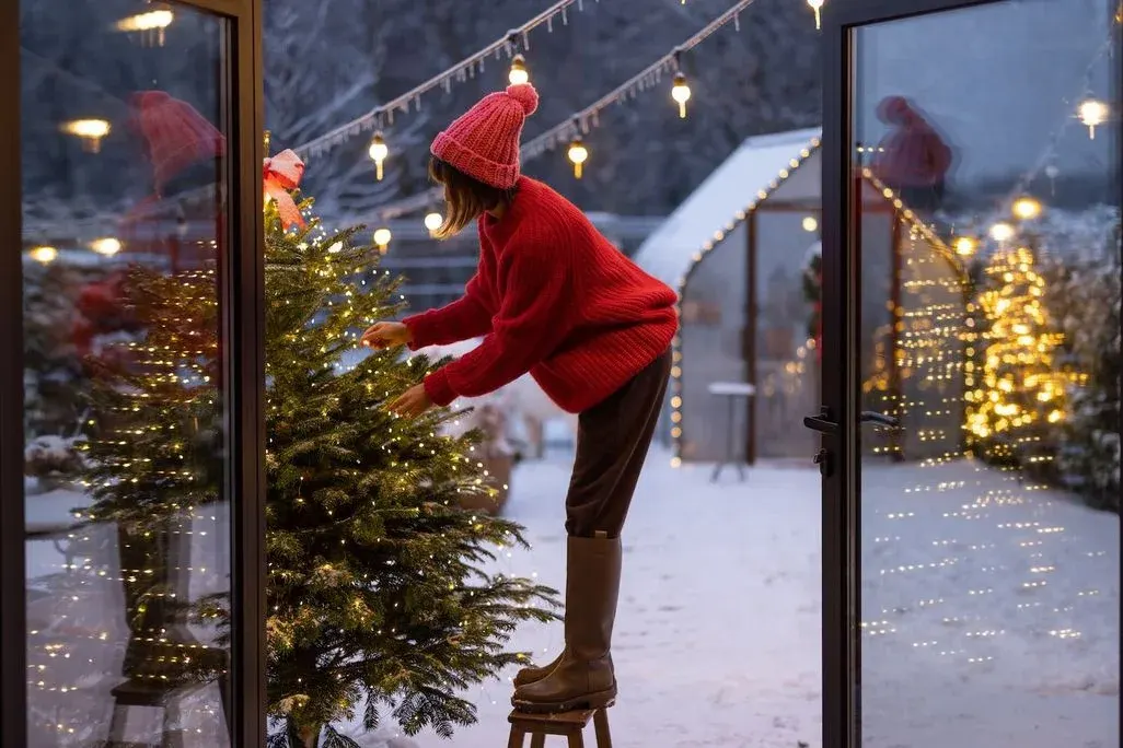 Eine Person in rotem Pullover und Mütze steht auf einem Holzhocker und schmückt einen grünen Weihnachtsbaum mit gelben Lichtern.