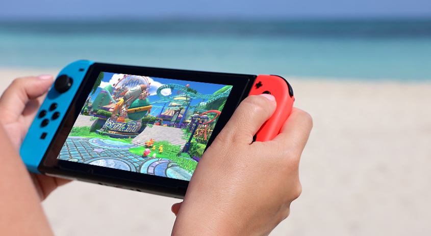 Eine Person hält eine Nintendo Switch mit einem Videospiel vor einem Strand mit blauem Wasser.