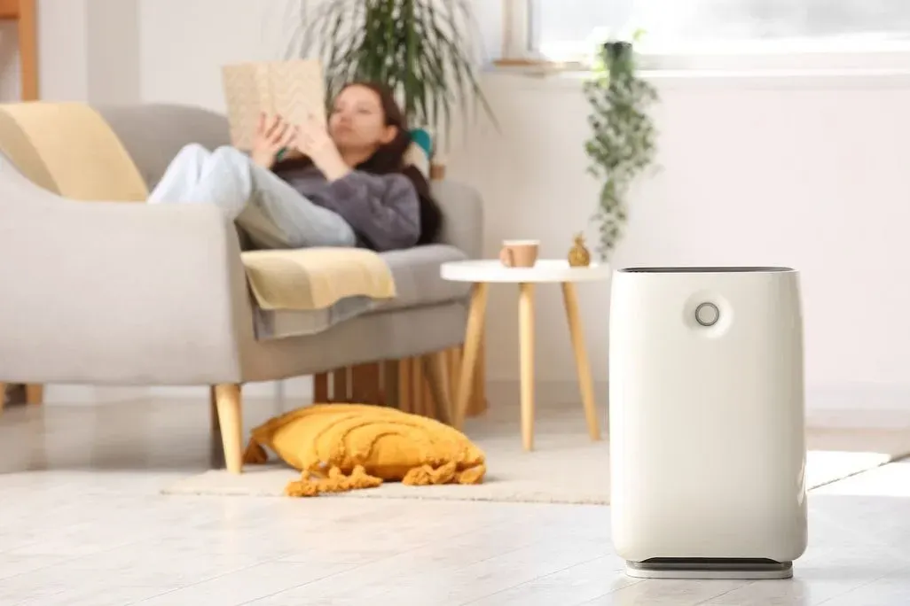In einem Wohnzimmer steht ein Luftreiniger im Vordergrund. Im Hintergrund ist etwas verschwommen eine Frau zu sehen, die auf der Couch liegt und liest.