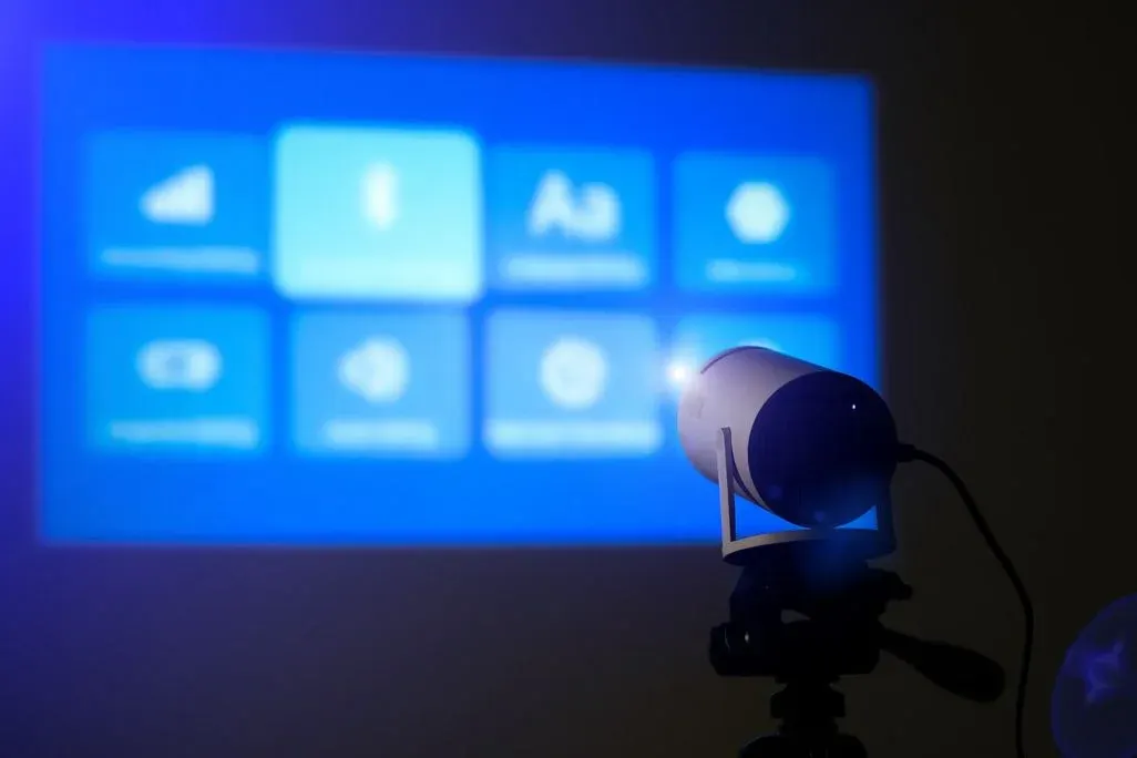 Kleiner Beamer auf einem Stativ projiziert ein blaues, unscharfes Menü mit Kachelsymbolen an eine Wand.