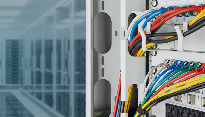 Auf der rechten Seite zeigt eine Nahaufnahme strukturierte, mehrfarbige Netzwerkkabel (rot, blau, gelb, grün, schwarz), die in ein Patchpanel innerhalb eines weißen Netzwerkschranks eingesteckt und sauber durch eine Kabelführung geleitet werden. Der linke Hintergrund zeigt verschwommen einen Gang in einem großen Serverraum, überlagert von einem blauen, digitalen Rastermuster.