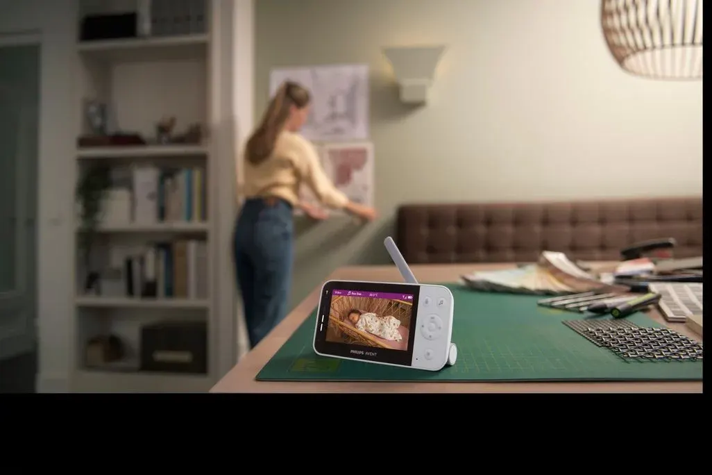 Elternteil arbeitet im Hintergrund an einem Schreibtisch, während im Vordergrund ein Babyphone mit Farbdisplay steht, auf dem ein schlafendes Baby zu sehen ist.