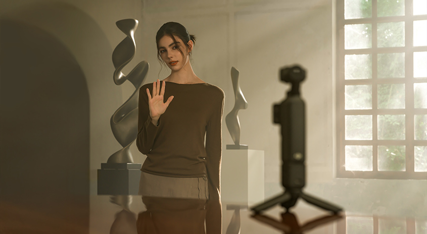 Frau in braunem Outfit in moderner Galerie winkt in Kamera. Hinter ihr abstrakte schwarze Skulpturen, große Fenster mit Tageslicht, minimalistisches Interieur.