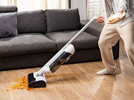 Jemand saugt mit einem Akkusauger Schmutz vom Boden auf, dahinter sieht man noch eine graue Couch.