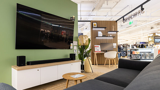 Einrichtung in einem Geschäft mit Fernseher, Lautsprechern, Soundbar auf weißem Schrank vor grüner Wand. Sitzgruppe im Vordergrund.