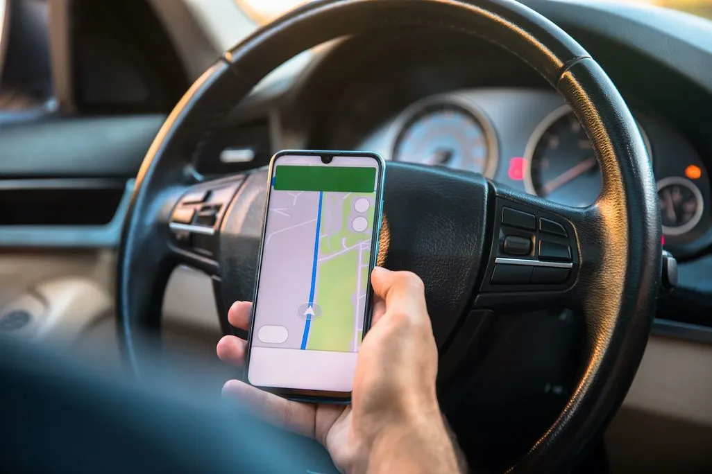 Eine Hand hält ein Smartphone mit Navigationsapp vor das Lenkrad eines Autos