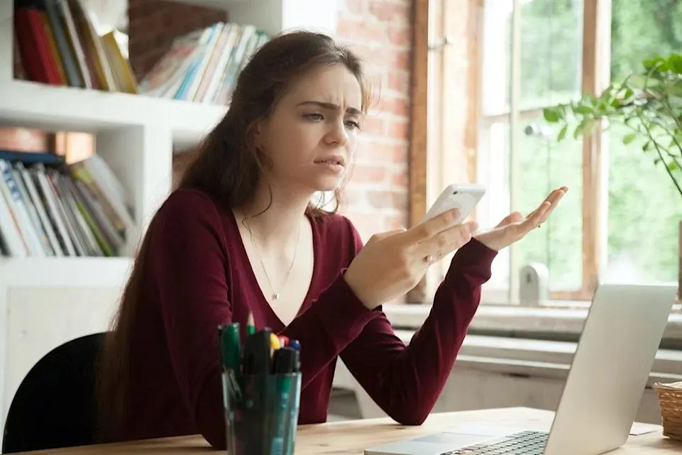 Eine Frau sitzt an einem Schreibtisch und schaut fragend auf ihr Smartphone.
