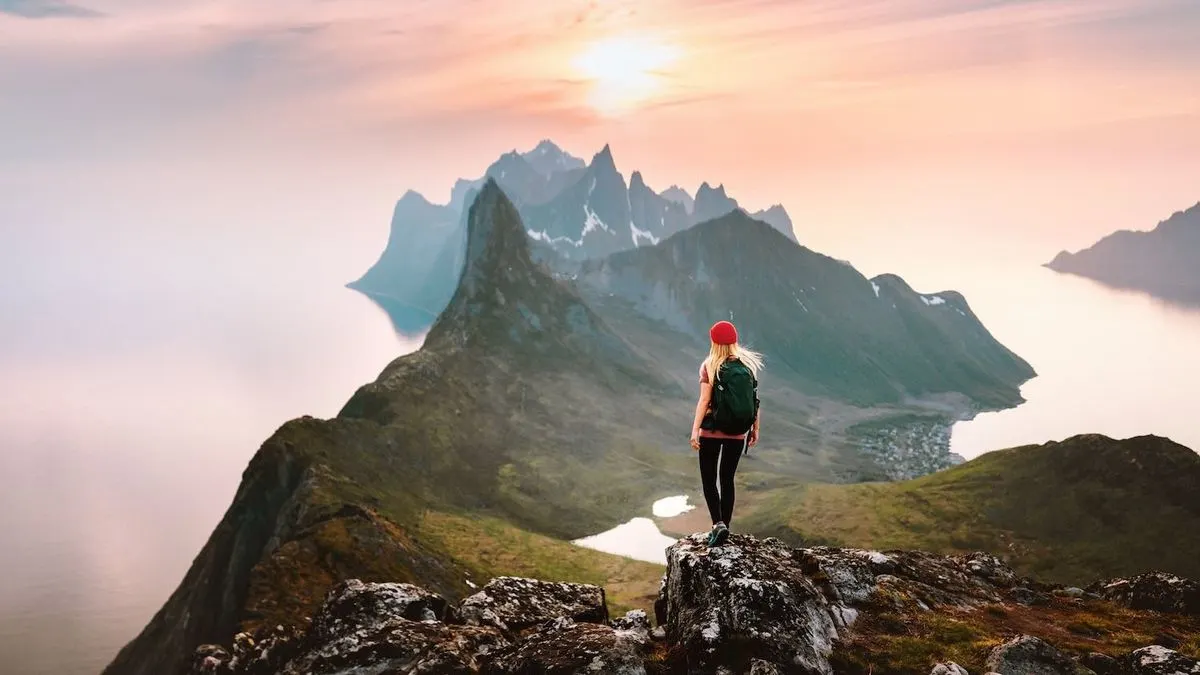 Person mit roter Mütze und Rucksack auf Felsen blickt auf grüne Berge, Fjord, Dorf und rosa-orangefarbenen Sonnenuntergang.