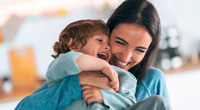Eine Person mit dunklen Haaren umarmt eine lachende Person mit braunen Haaren vor einem hellen, unscharfen Hintergrund.