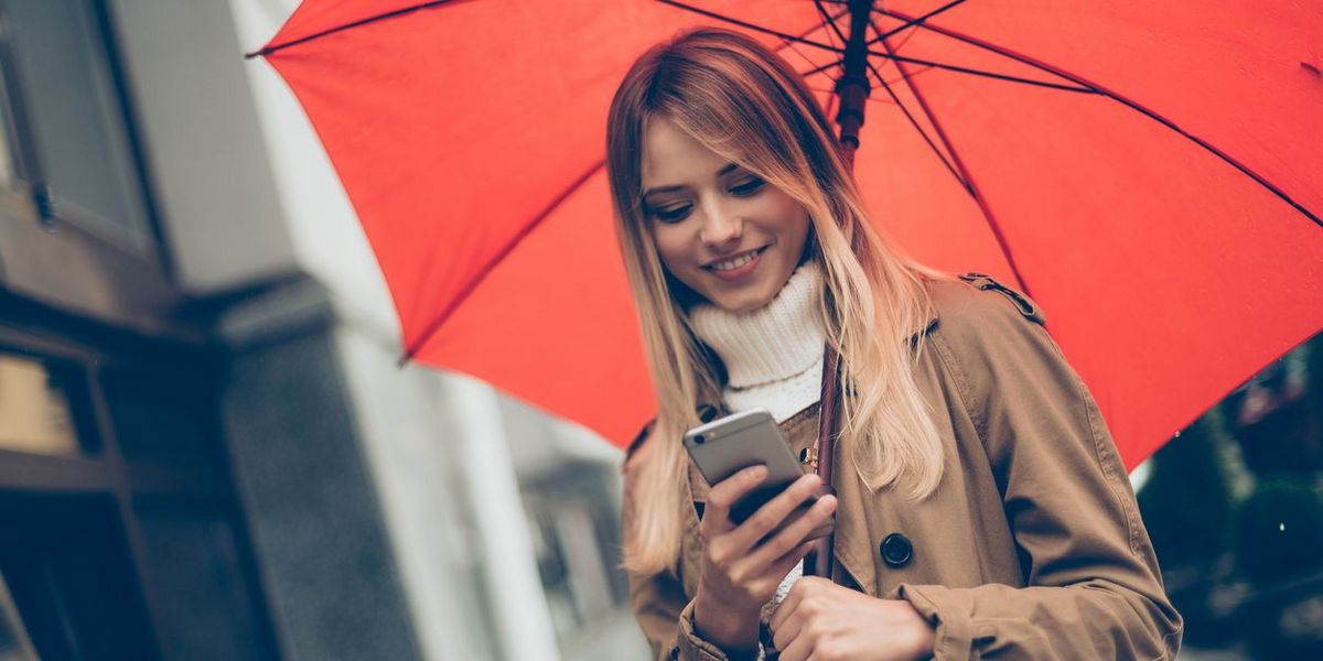Person in braunem Mantel und weißem Pullover blickt auf ein Smartphone, hält einen roten Regenschirm. Unscharfe Gebäude im Hintergrund.