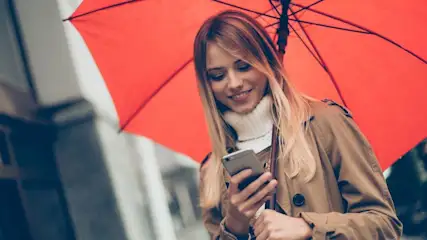 Person in braunem Mantel und weißem Pullover blickt auf ein Smartphone, hält einen roten Regenschirm. Unscharfe Gebäude im Hintergrund.