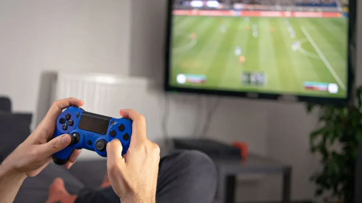 Hände halten einen blauen Videospiel-Controller mit schwarzen Knöpfen. Eine Person sitzt mit dunkler Hose und orangefarbenen Socken.