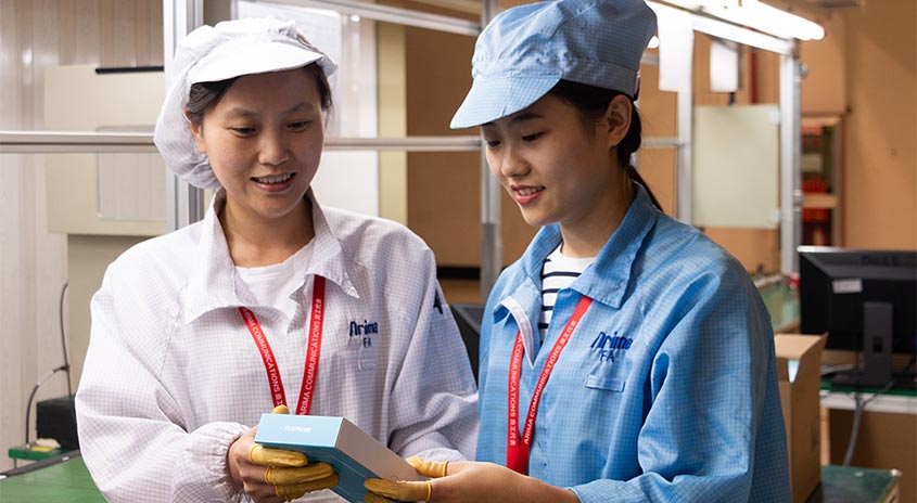 Zwei Personen in Arbeitskleidung und Hauben betrachten eine hellblaue Schachtel in einer Fabrik mit Computern.