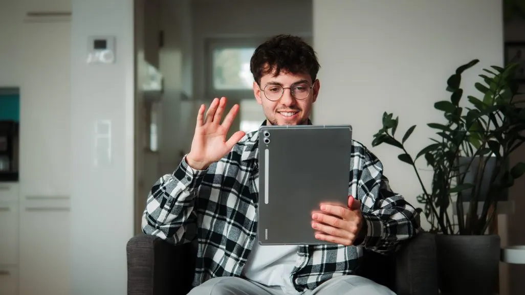 Lächelnder Mann mit Brille winkt in einem Videocall während er ein Tablet hält