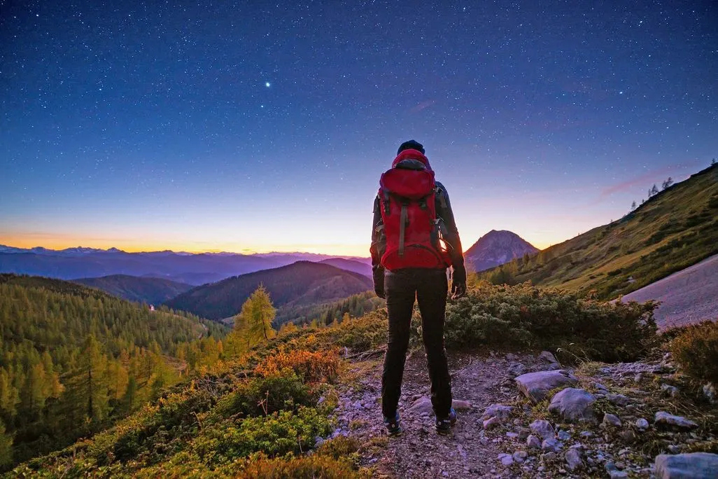 Eine Person mit rotem Rucksack steht auf einem Berg und blickt in die Sterne