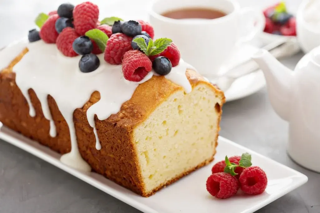 Rührkuchen mit weißem Zuckerguss, Himbeeren, Heidelbeeren und Minzblättern auf weißer Platte. Im Hintergrund Teetasse und Kanne.