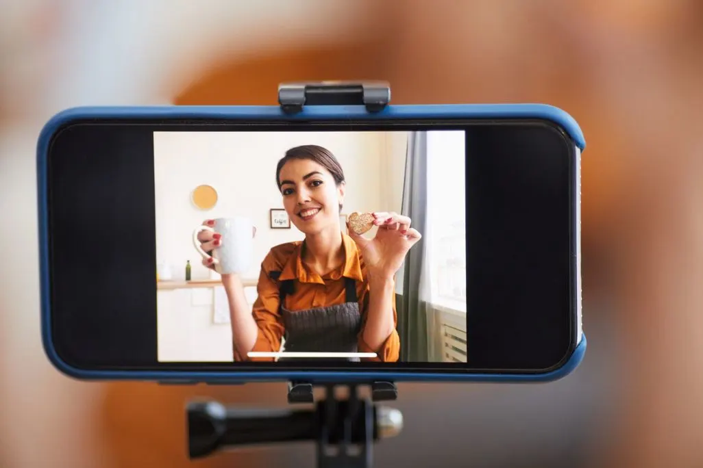 Frau filmt sich selbst mit einem Smartphone und hält eine Tasse und einen Keks in die Kamera.