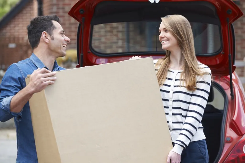 Zwei Personen heben gemeinsam einen großen Karton aus dem Kofferraum eines roten Autos