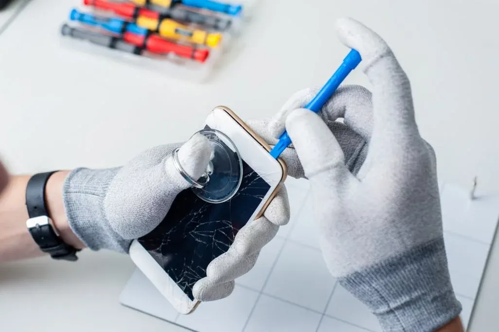 Hände in hellgrauen Handschuhen öffnen ein weißes Smartphone mit gesprungenem Bildschirm mittels Saugnapf und blauem Hebelwerkzeug.
