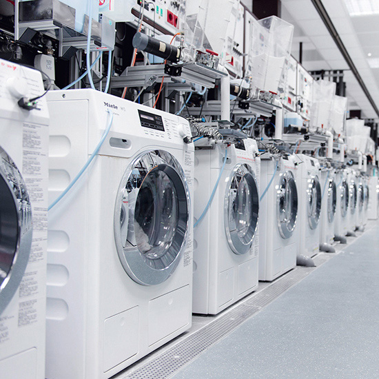 Reihe weißer Waschmaschinen mit Chromakzenten unter technischen Geräten in hellem Ausstellungsraum.