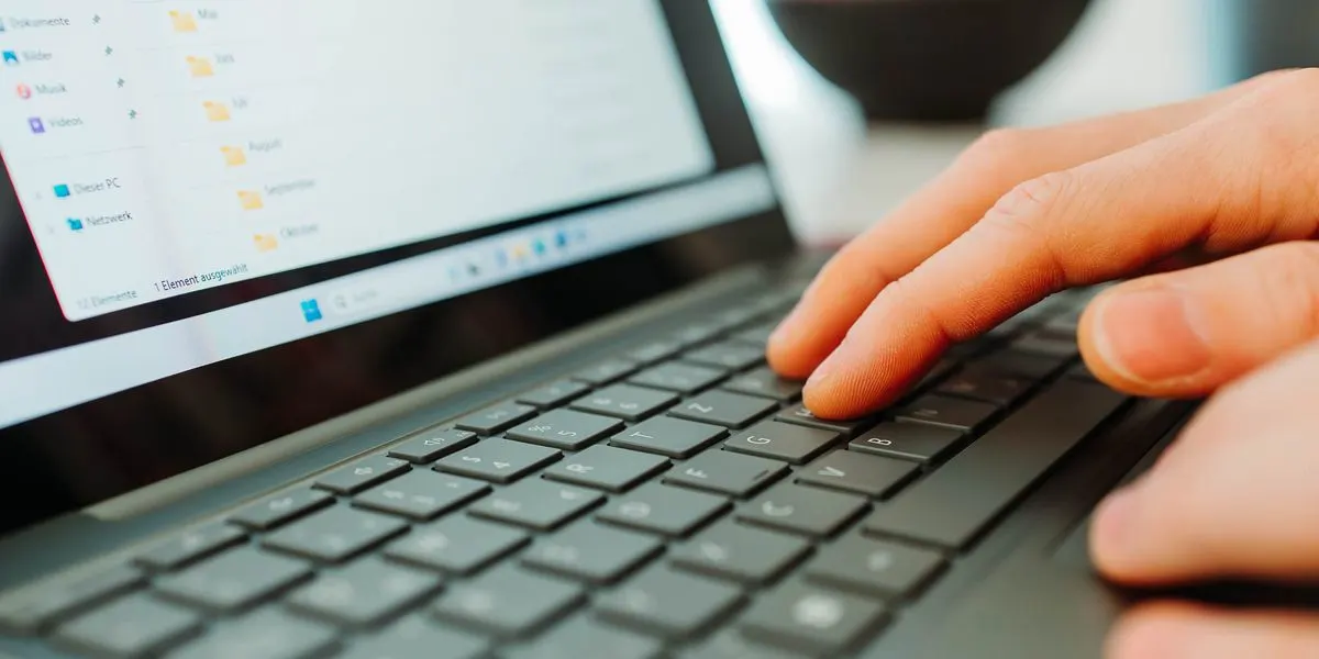 Nahaufnahme einer Hand auf einer dunkelgrauen Laptop-Tastatur, Finger tippen. Der Bildschirm zeigt Dateiverzeichnisse.