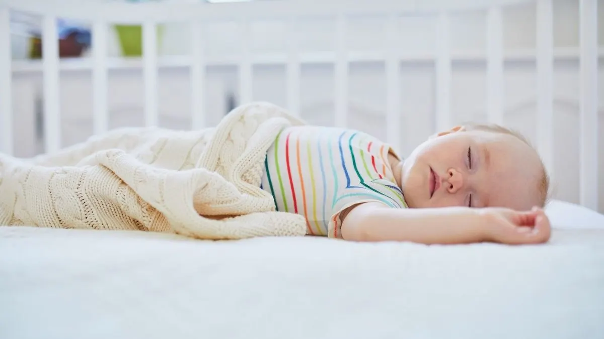 Baby mit gestreiftem Body schläft auf dem Rücken in einem weißen Gitterbett und ist mit einer hellen Strickdecke zugedeckt.